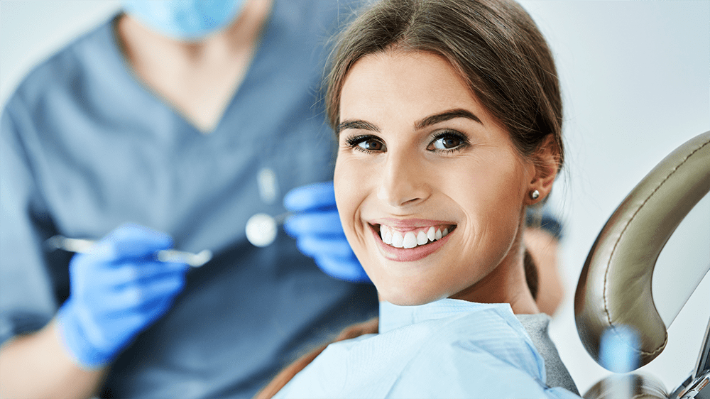 A woman smiling brightly in a dental chair with a dentist blurred in the background