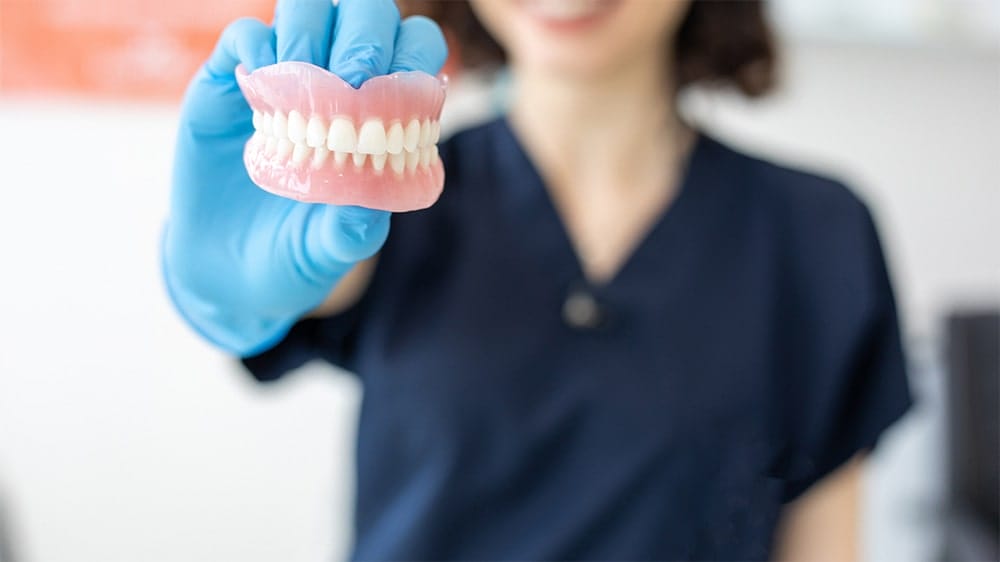 A dental assistant wearing gloves holding a full set of dentures toward the camera
