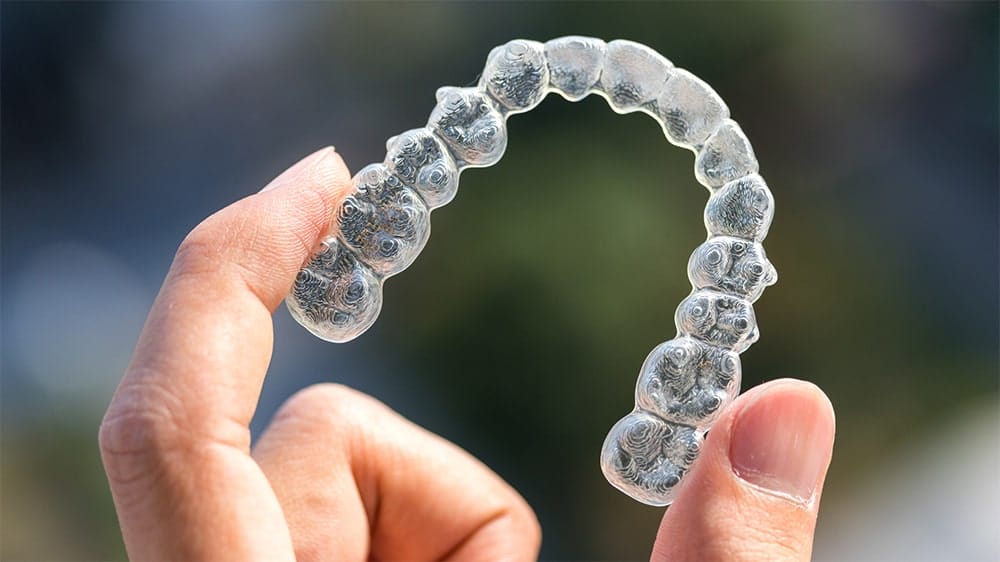 Close-up of a clear aligner held between two fingers against a blurred background