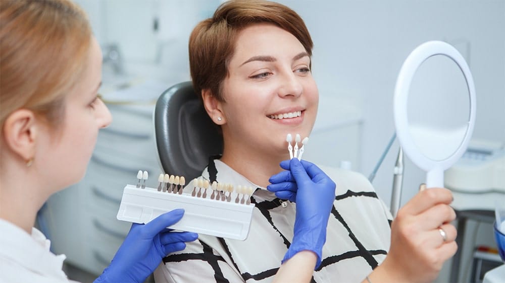 A woman holding a mirror and selecting a tooth shade from a dental shade guide held by a dentist