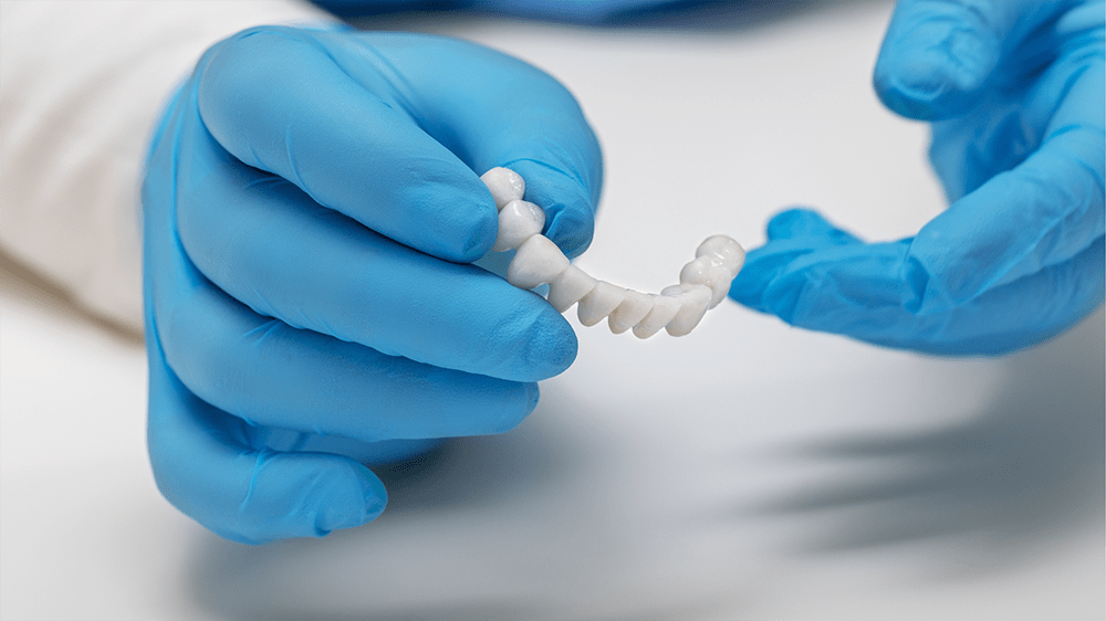 Gloved hands holding a dental bridge model over a clean white surface