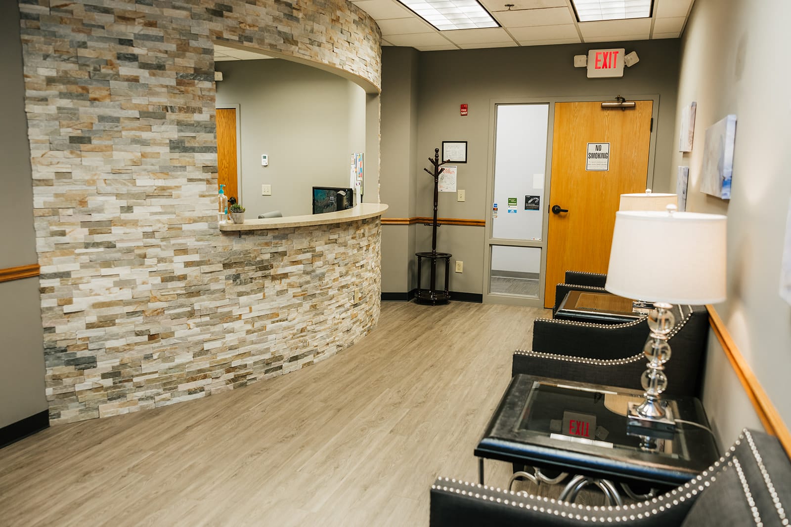 Reception area of a dental office with a curved stone-front desk, wood flooring, waiting chairs, and hallway leading to treatment rooms.