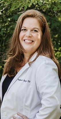 Dr. Stephanie, DDS, wearing a white coat smiling outdoors in front of greenery