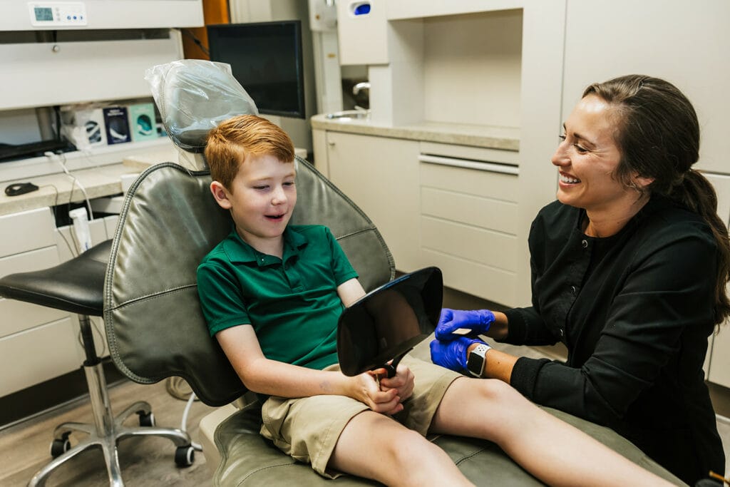 Young boy smiling and holding a mirror while sitting in a dental chair