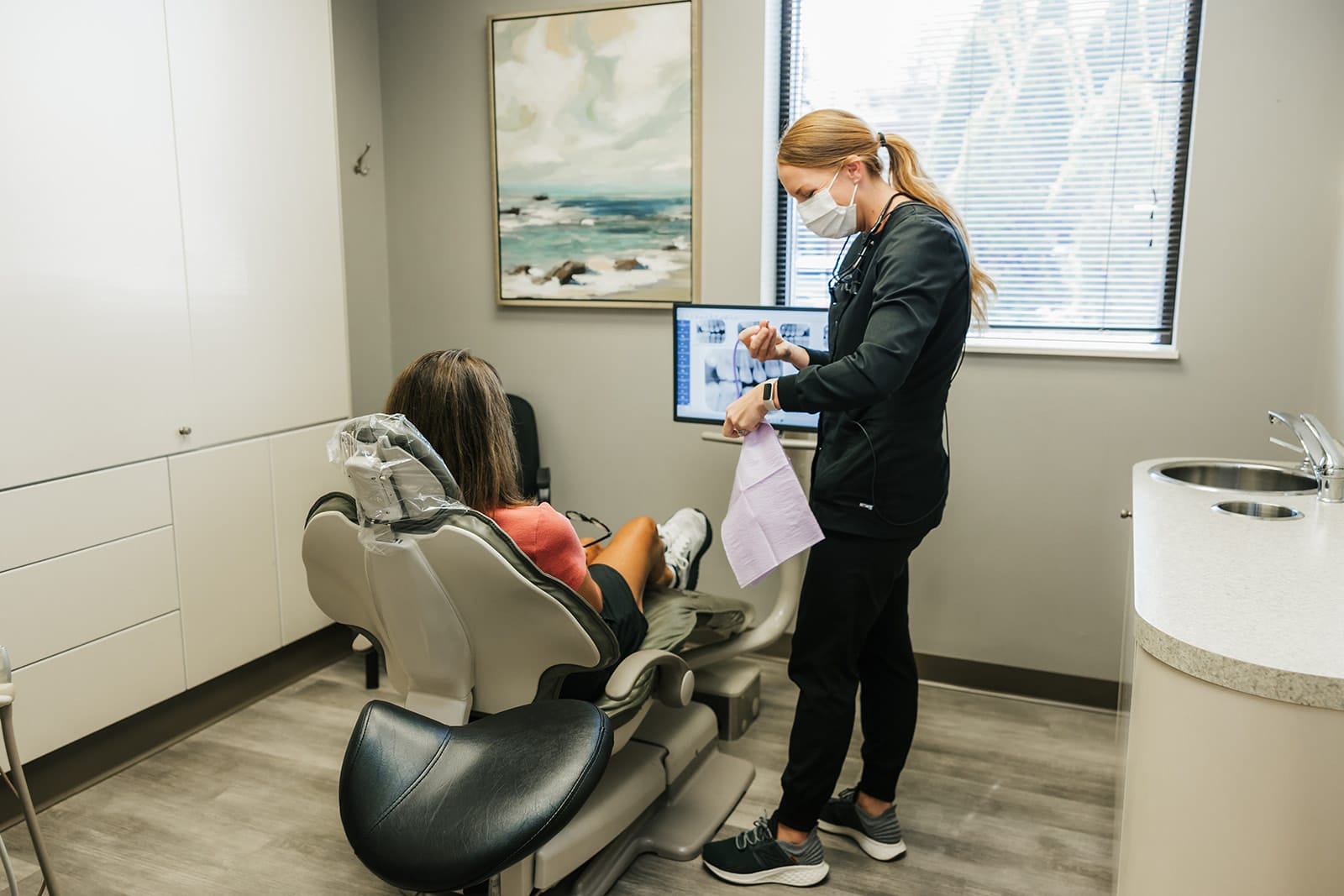 Dentist wearing a mask reviewing X-rays and paperwork with a patient seated in a dental chair during an exam.