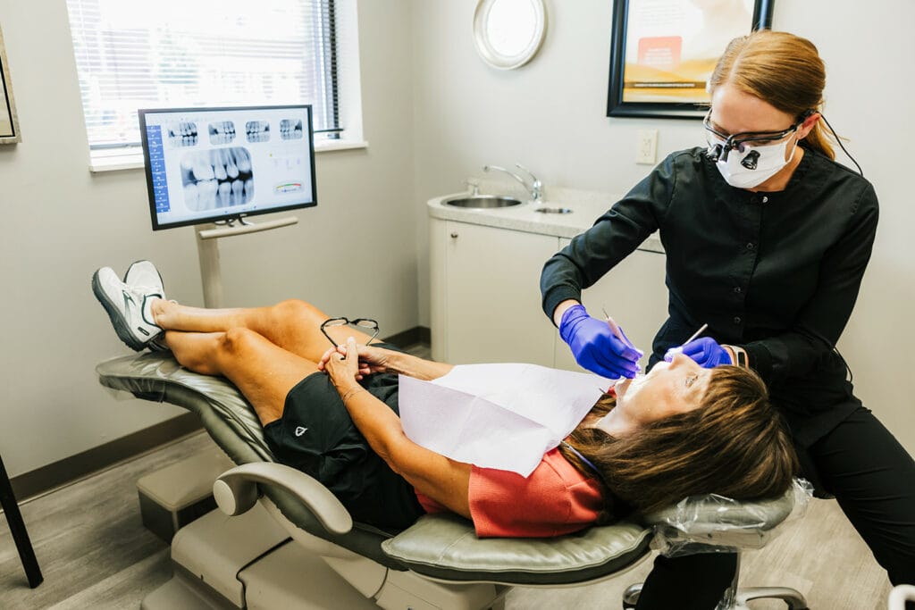 A dentist in protective gear working on a female patient reclining in a dental chair with an X-ray screen visible