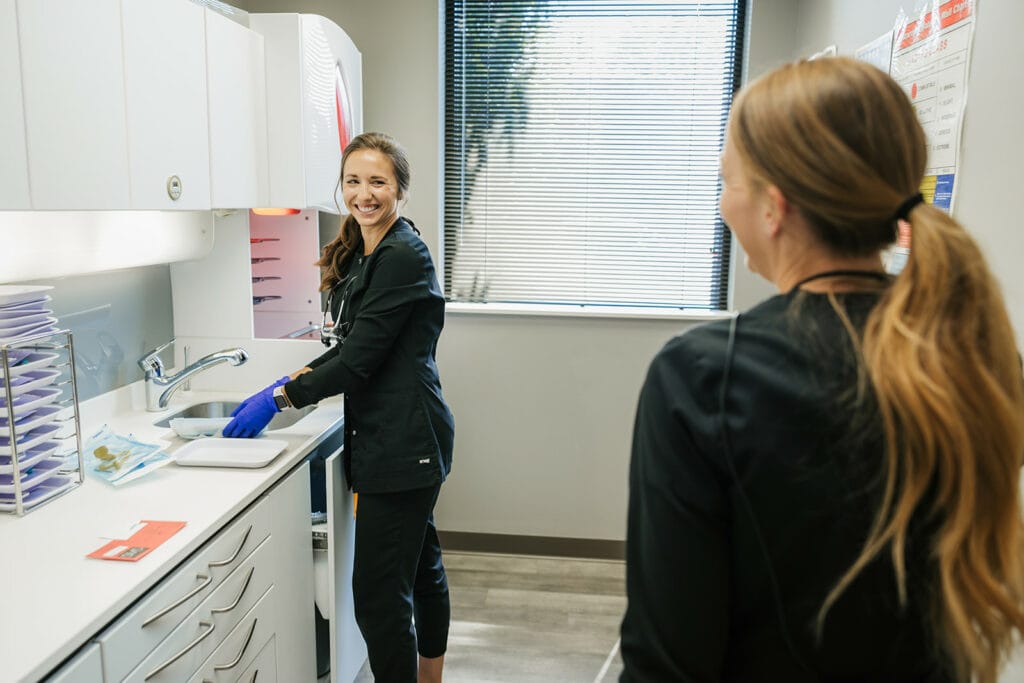 A dental assistant smiling while preparing tools near a sink as a colleague looks on