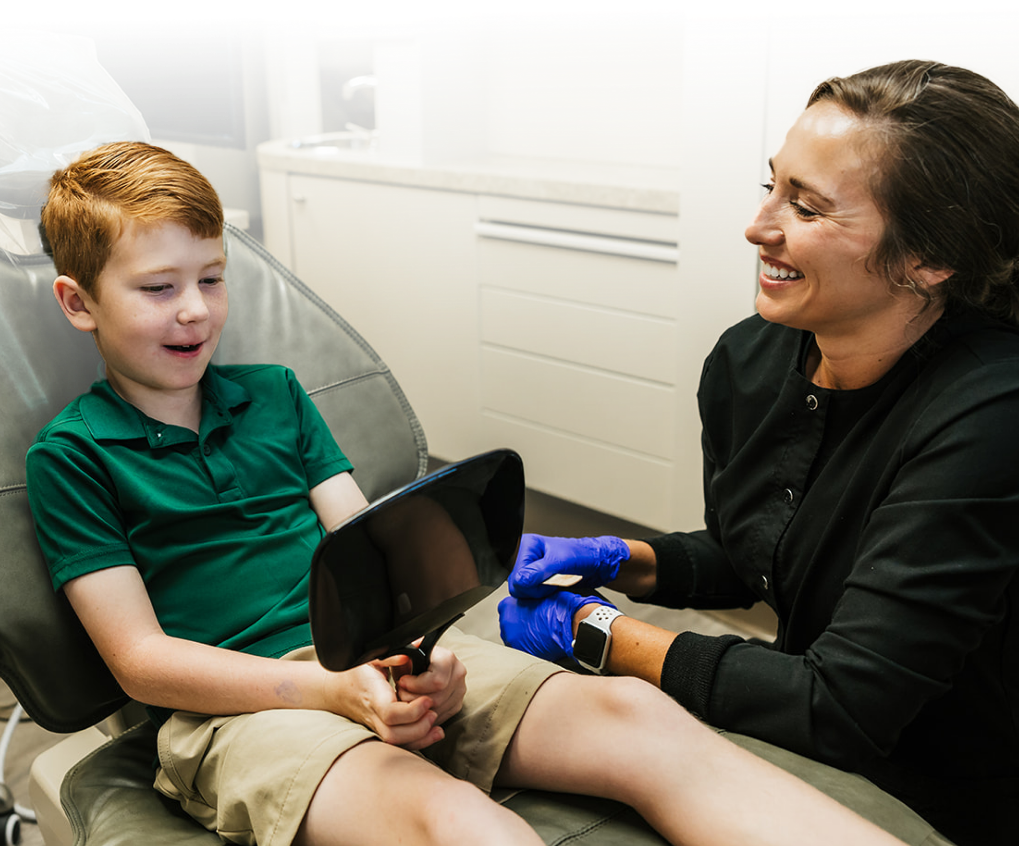 A young patient in a dental chair using a handheld mirror while a smiling dental professional explains preventive oral hygiene at Stephanie Briggs, DDS.