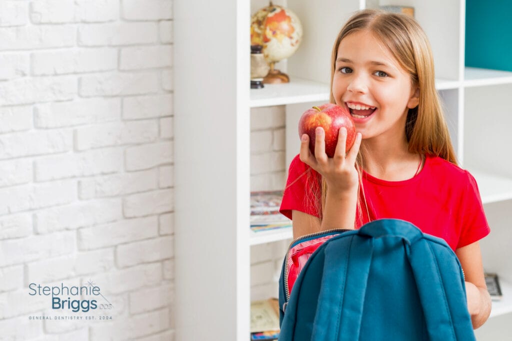 Smiling young girl in a red shirt holding an apple and a blue backpack, standing in a bright room with a white brick wall and bookshelf.
