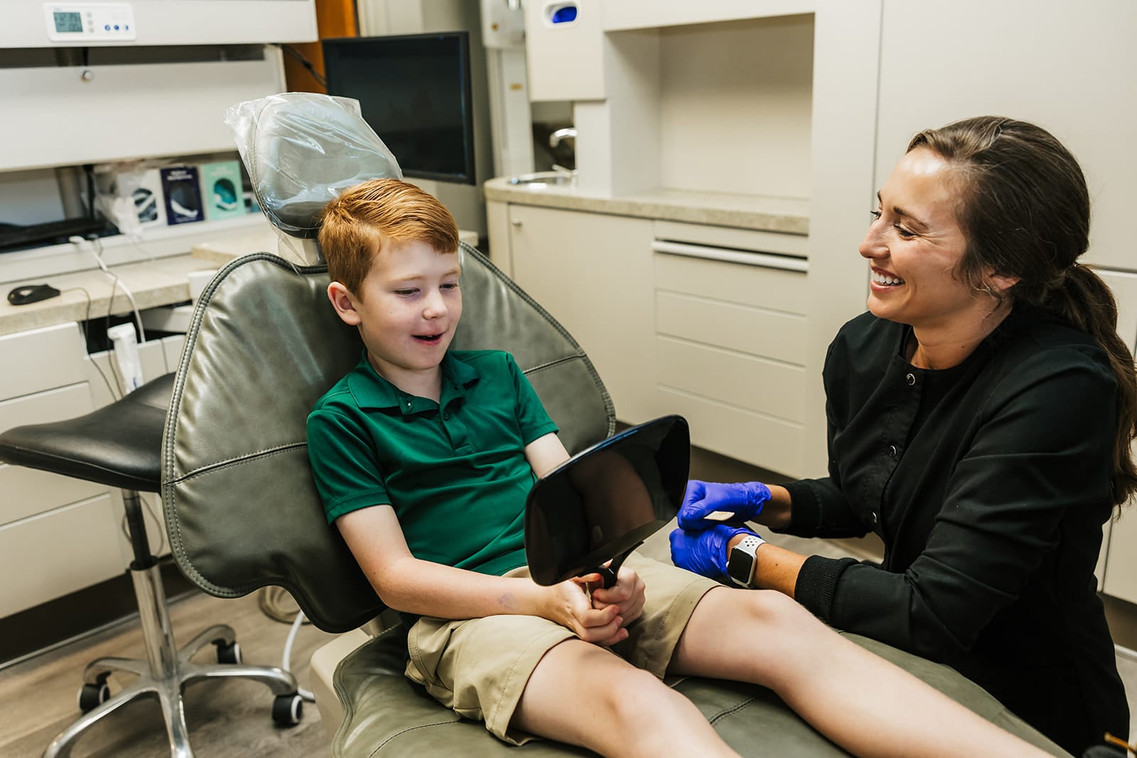 Young boy sitting on a dental chair holding a mirror with Dr. Stephanie Briggs